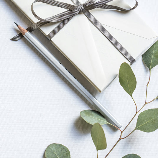“Flat lay of a neatly wrapped envelope tied with a ribbon, a pencil, and sprigs of greenery on a white background — evoking simplicity, care, and thoughtful gifting.”