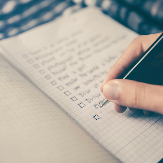 “Close-up of a hand writing a checklist in a notebook, reflecting the pressure of modern to-do lists and decision fatigue.”
