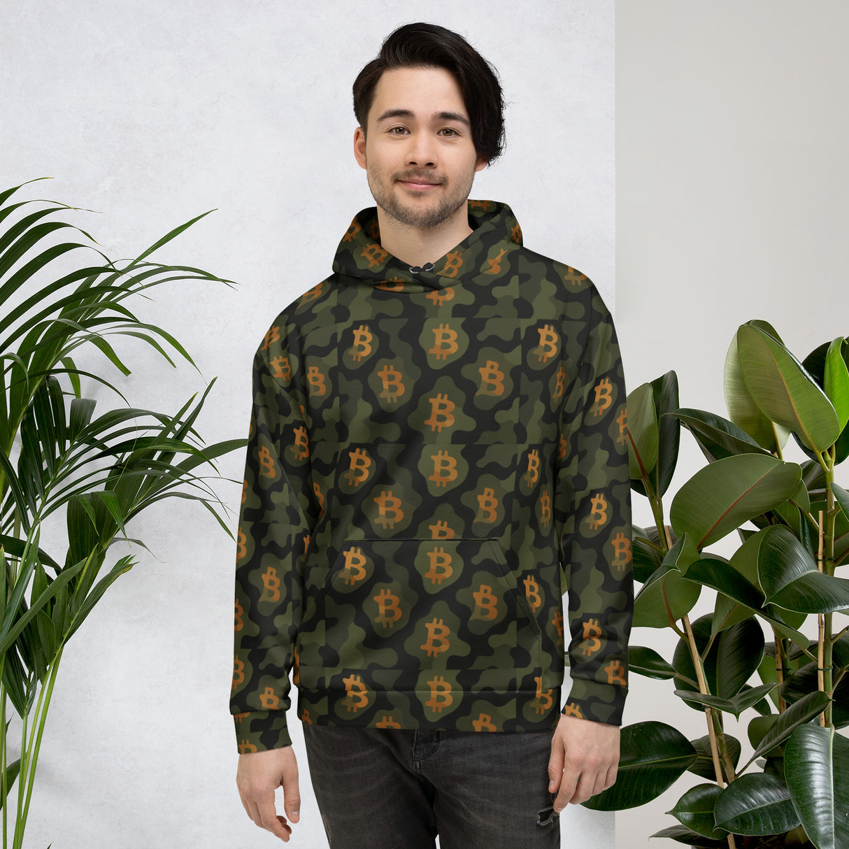 Person wearing a green camouflage hoodie with Bitcoin symbols, standing between two plants.