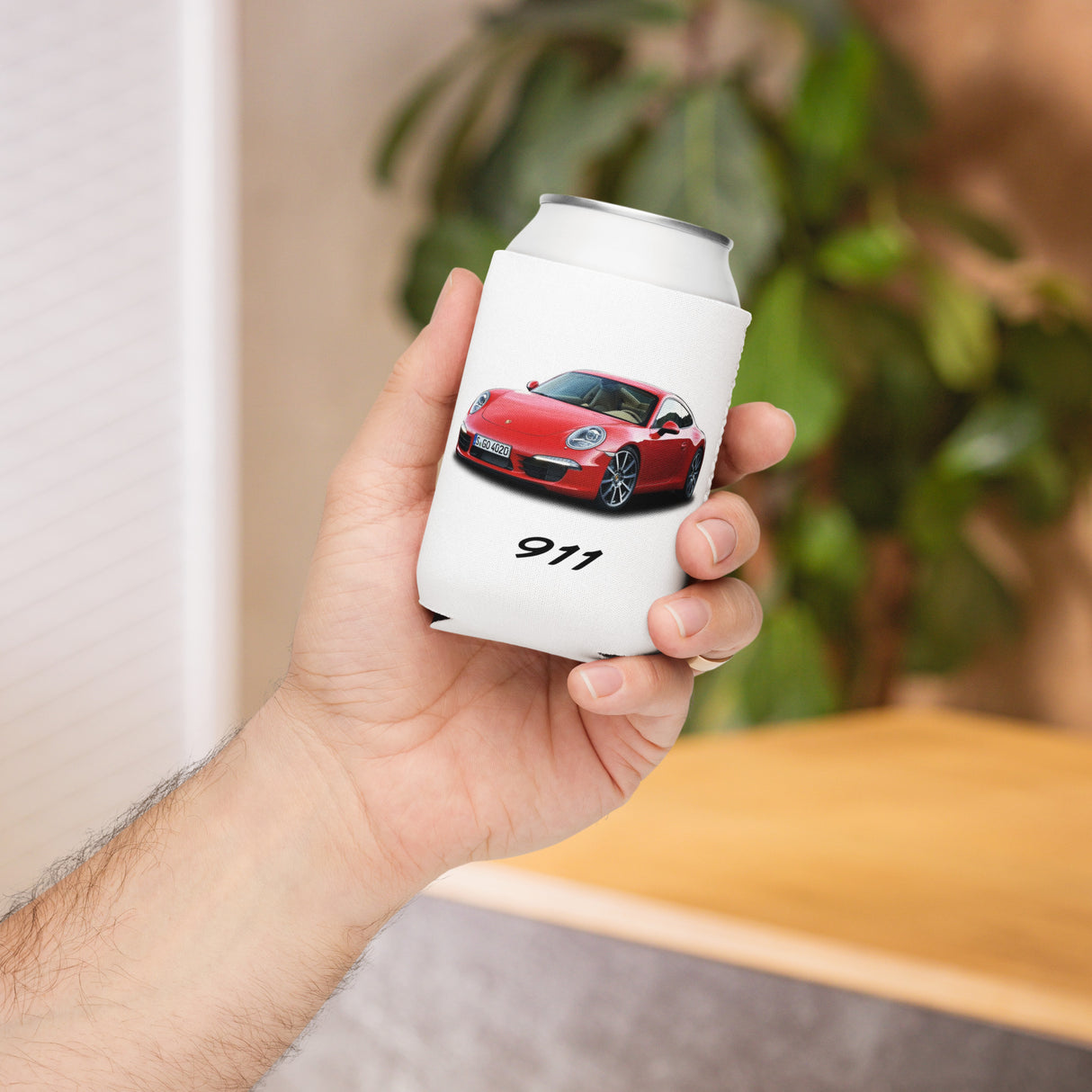 Hand holding a white can cooler with a red car design and '911' text, blurred indoor background