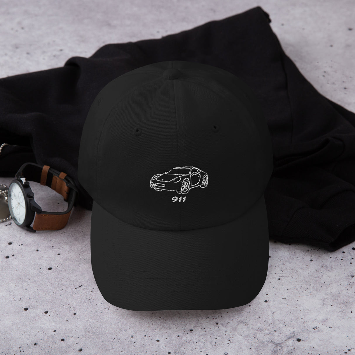 Black low-profile dad cap with white embroidered Porsche outline design displayed on desk next to sunglasses and keys