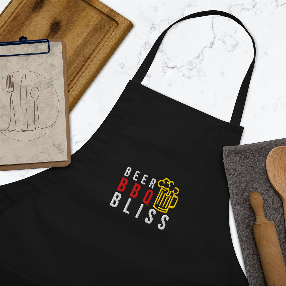 Black apron with 'Beer BBQ Bliss' design on a marble surface with kitchen items.