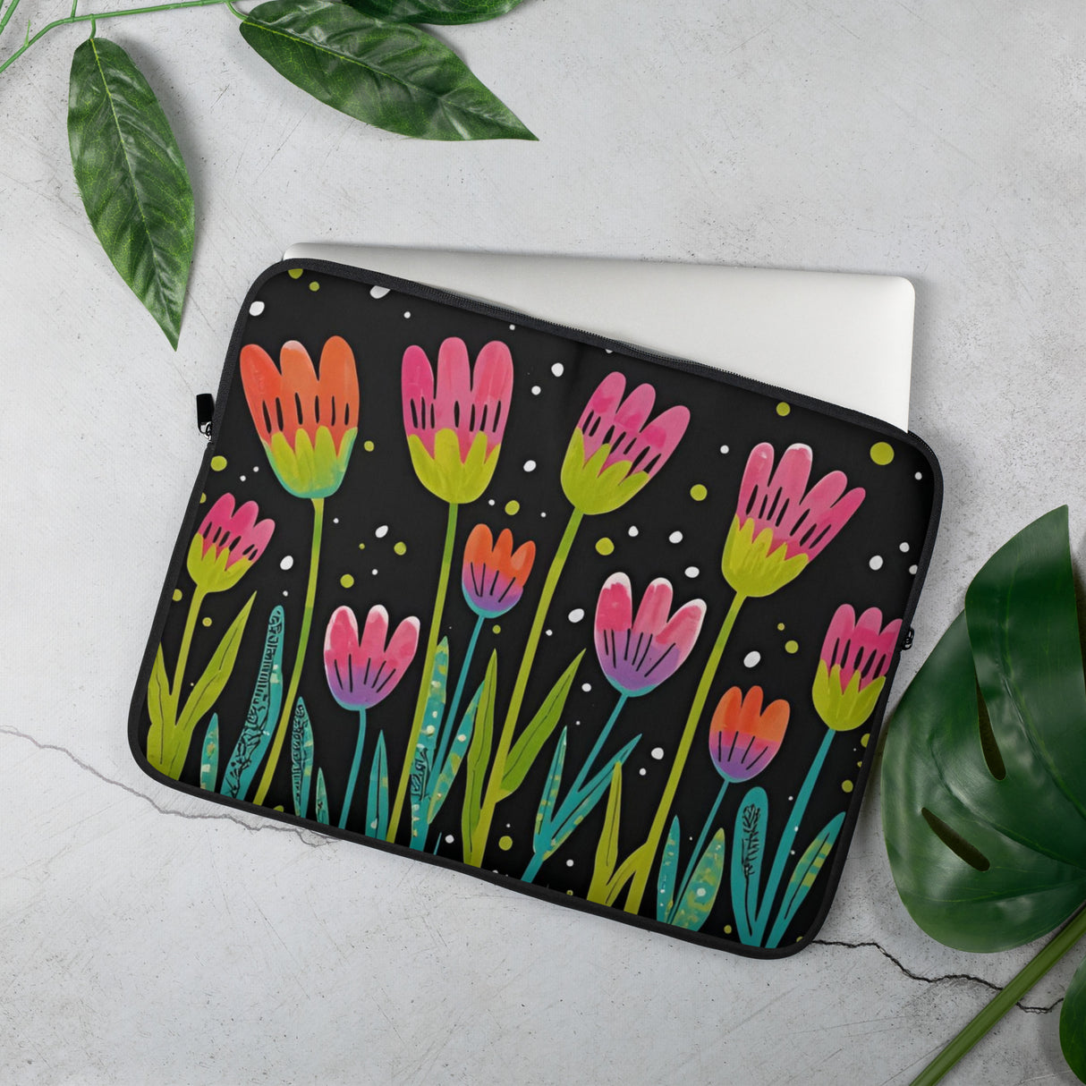 Black laptop sleeve with colorful floral design, styled flat lay with greenery.