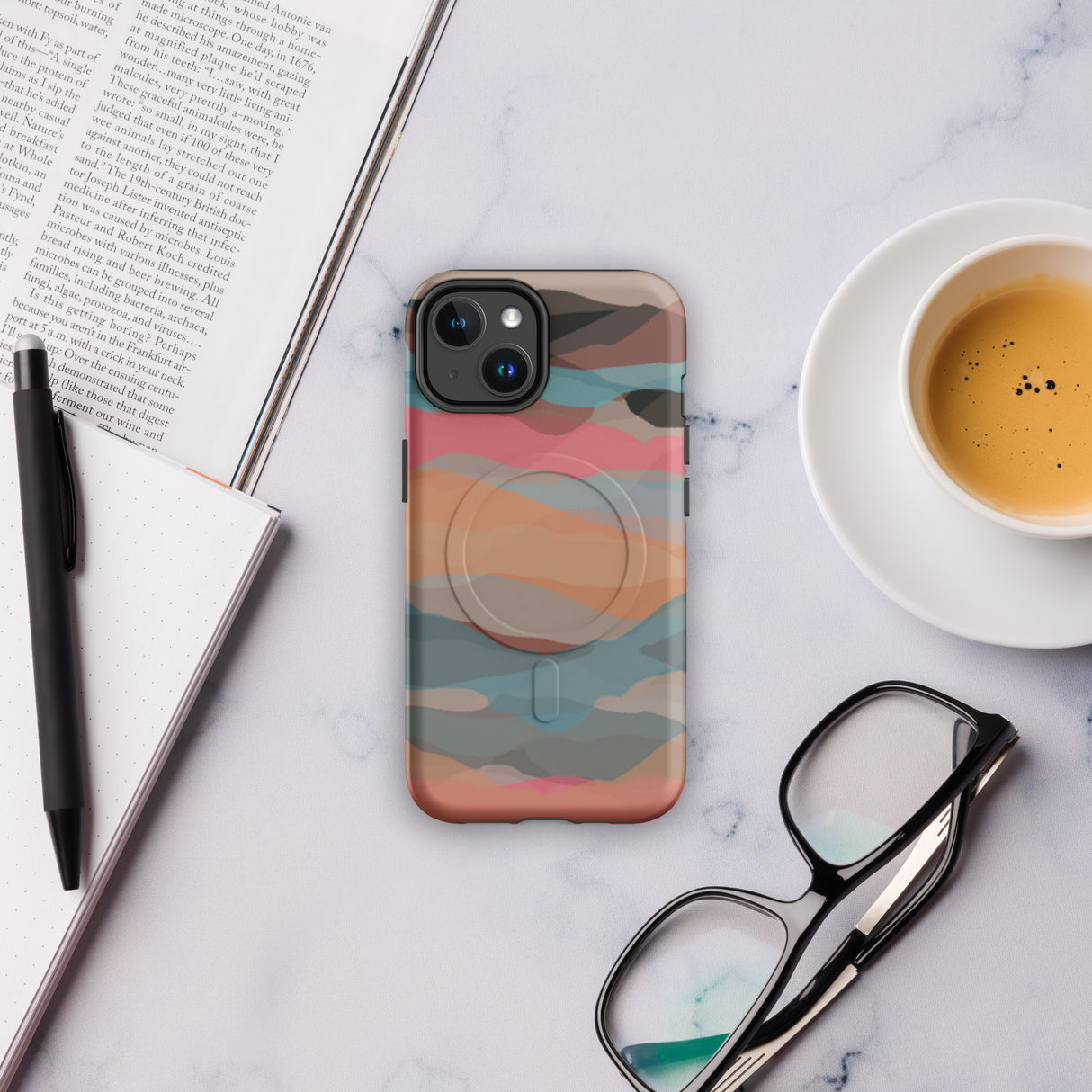 Phone case with colorful design on a marble surface with a book, pen, coffee cup, and glasses.